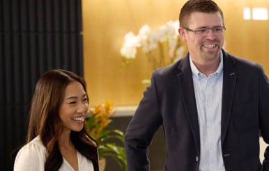 A woman and a man are smiling in a well-lit indoor setting. The woman has long dark hair and is wearing a white top. The man is wearing glasses and a dark blazer. A plant with white flowers is visible in the background.