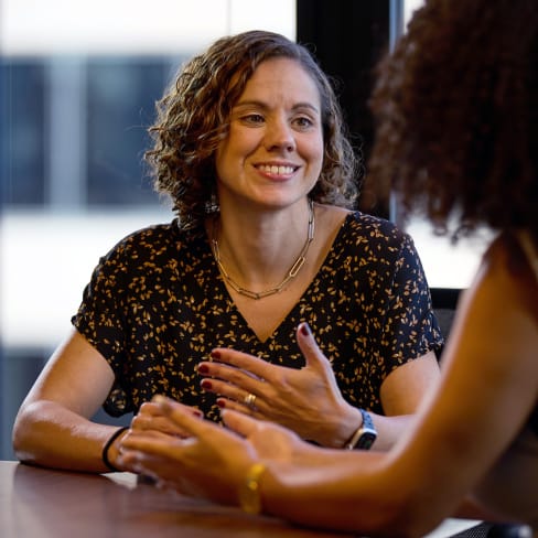 A woman with curly hair, wearing a patterned shirt and necklace, smiles as she gestures with her hands in a conversation with another person. The setting appears to be an office or a meeting room.