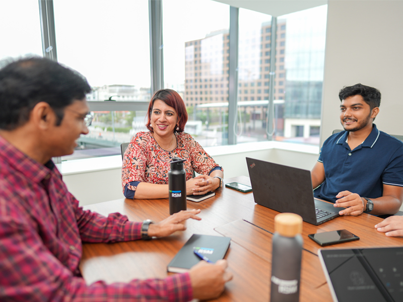 Three people sit around a conference table in a modern office, engaged in discussion. Two have laptops open, and all appear focused and smiling. Large windows show buildings outside in the background.
