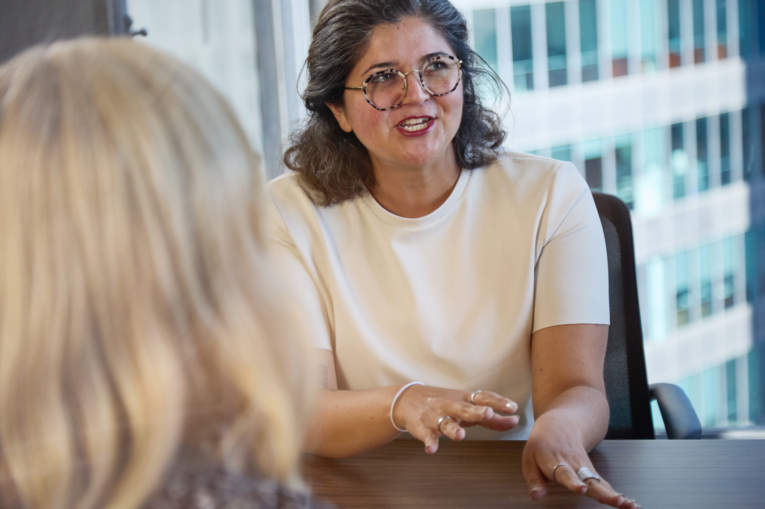 A woman with glasses and gray hair, wearing a white top, gestures with her hands while speaking to another person in an office setting with large windows and tall buildings outside.