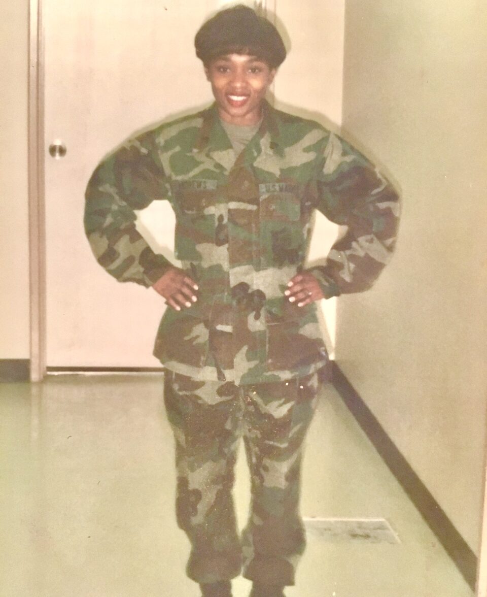 A person wearing a camouflage military uniform and beret stands indoors, smiling with hands on hips, against a beige wall and a closed door in the background.