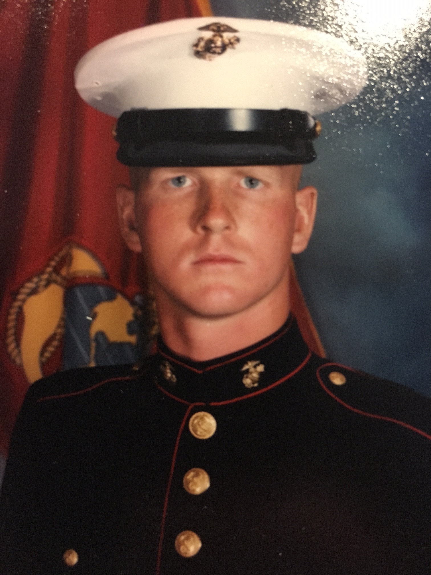 A young Marine in dress uniform and white cap looks straight ahead, standing in front of a US Marine Corps flag with the emblem visible in the background.