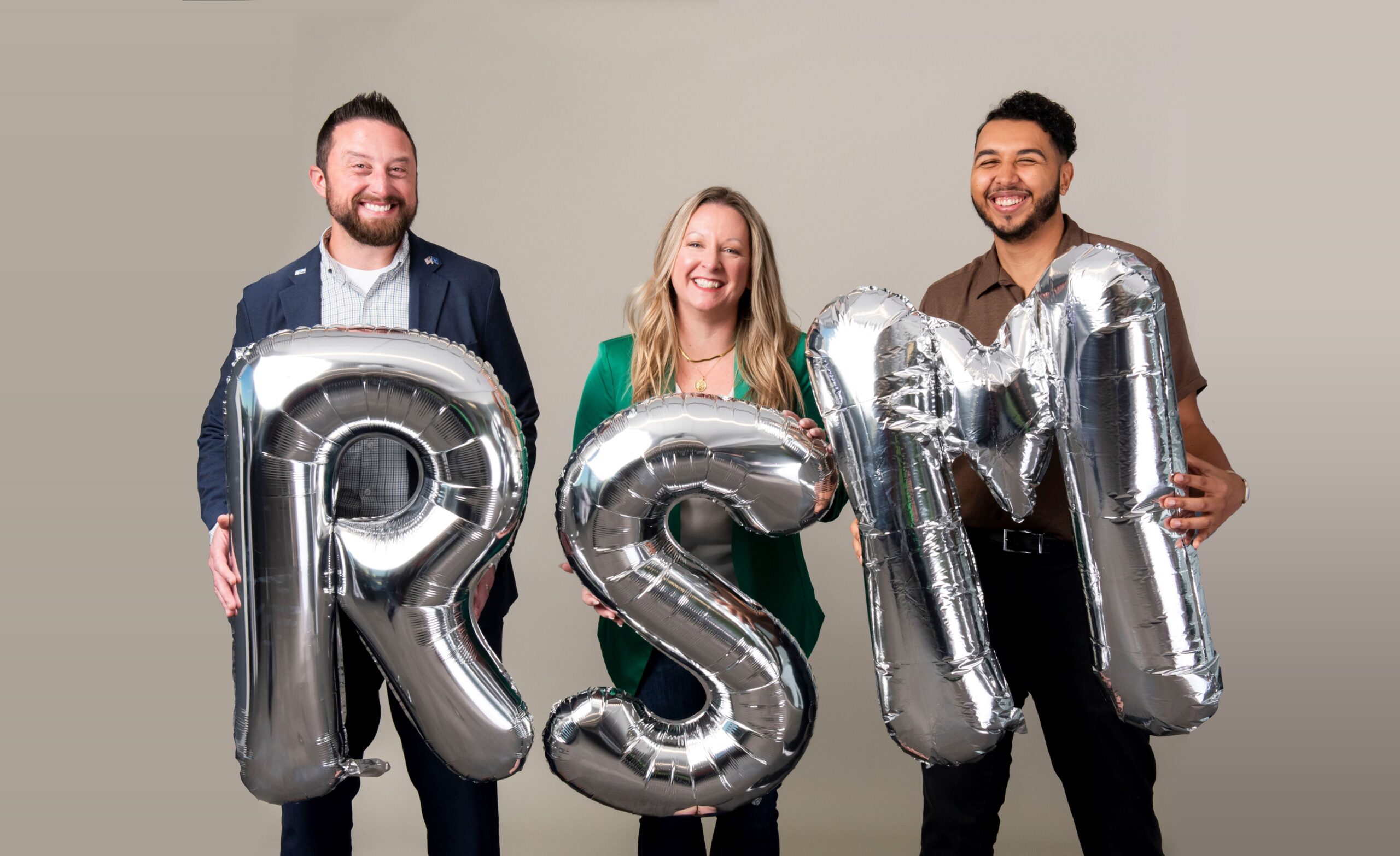 Three people stand smiling, each holding a large silver balloon shaped like the letters "R", "S", and "M" against a neutral background.