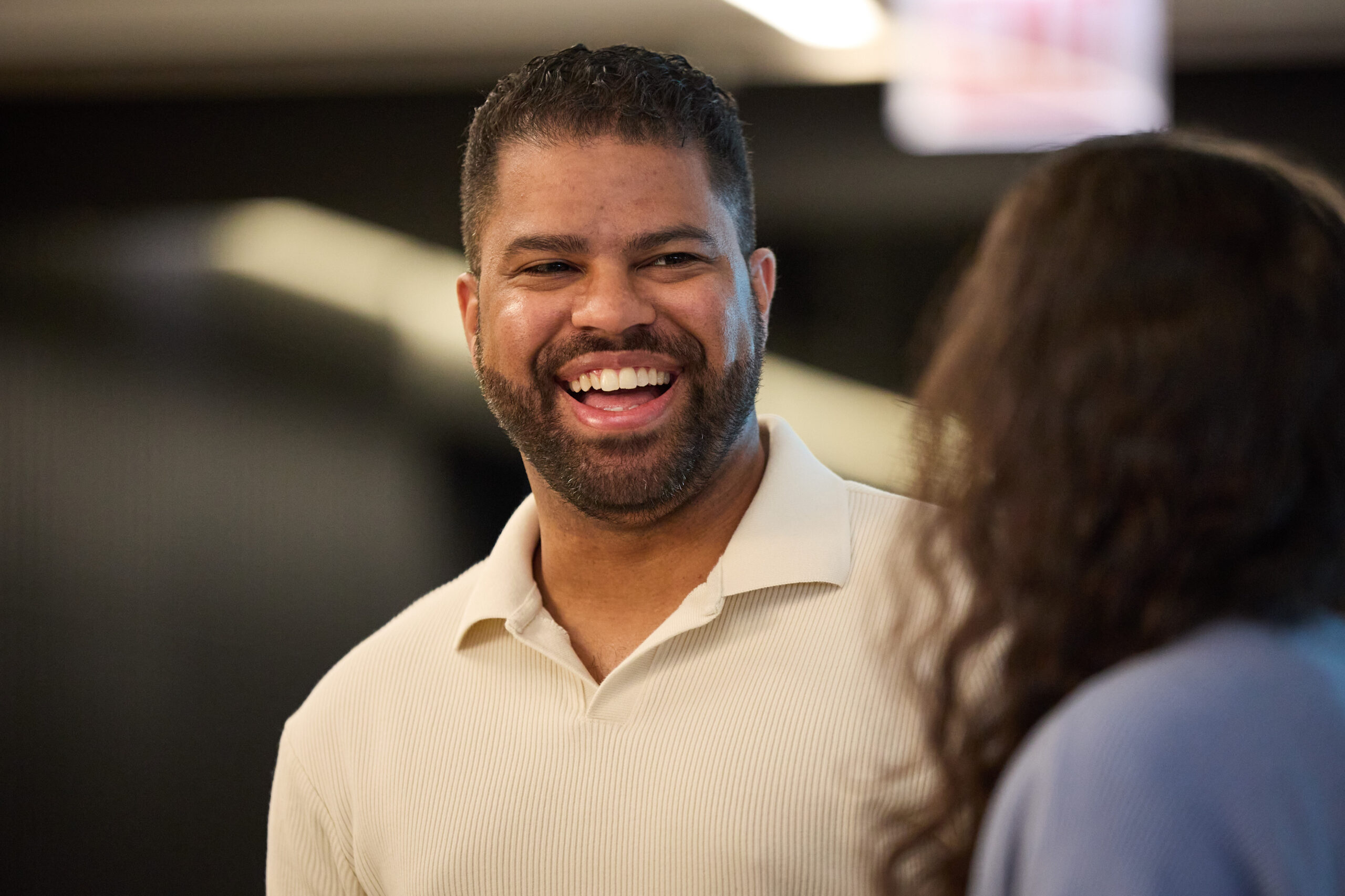 A man with a beard wearing a light-colored polo shirt smiles and laughs while talking to a person with long hair indoors. The background is blurred.