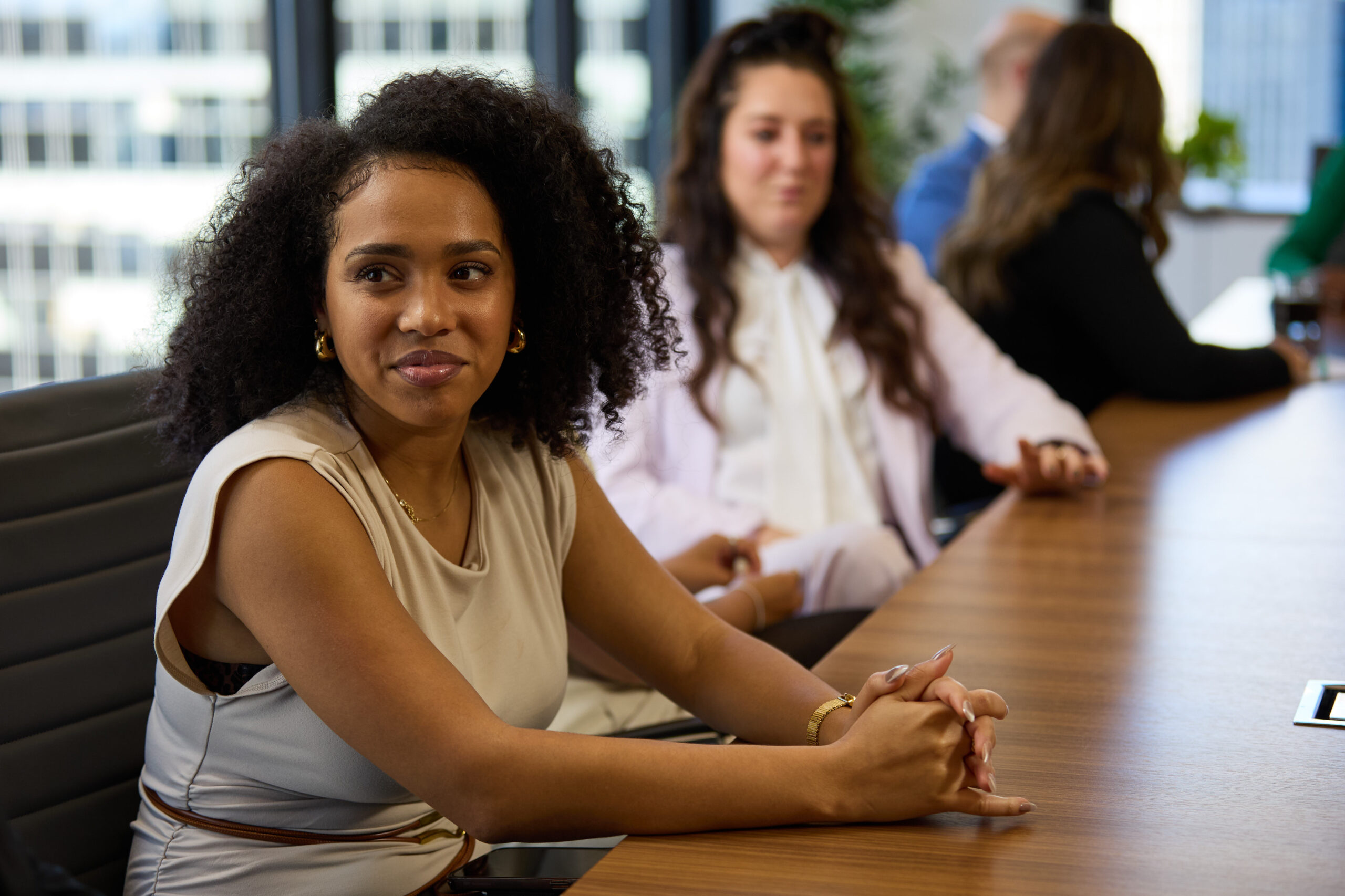 A woman with curly hair sits at a conference table, smiling and looking to the side. Two other people are seated beside her, slightly out of focus, in a modern office setting.