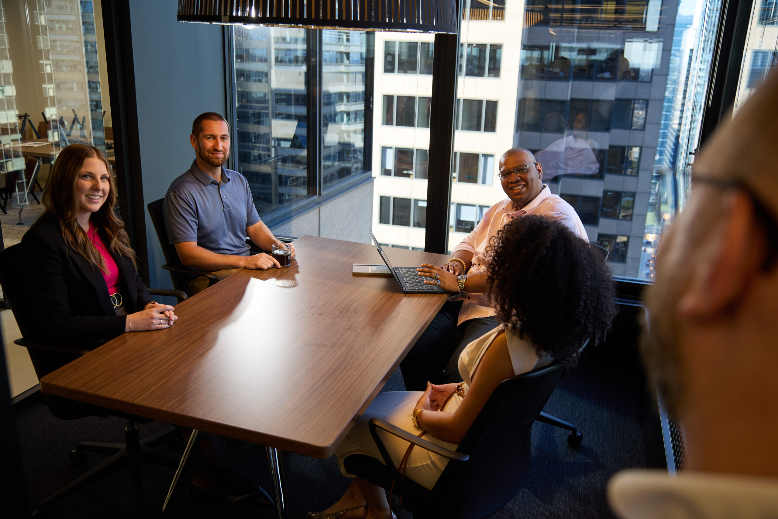 Five people sit around a conference table in a modern office with large windows overlooking city buildings, engaged in a meeting. Some are smiling, and one person uses a laptop.