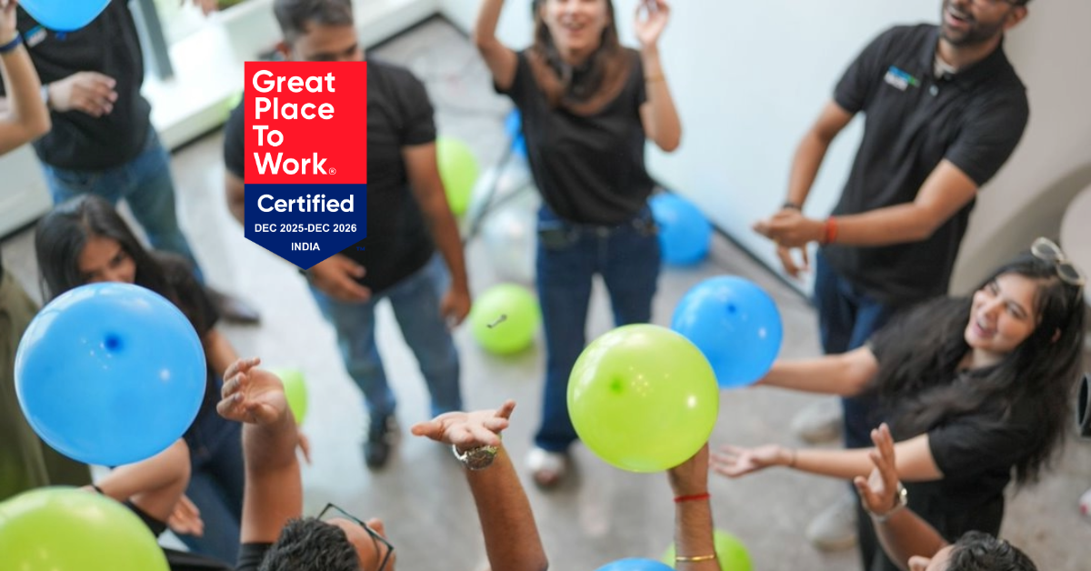 A group of people smiling and playing with blue and green balloons indoors. The “Great Place To Work Certified” badge is visible on the left side of the image.