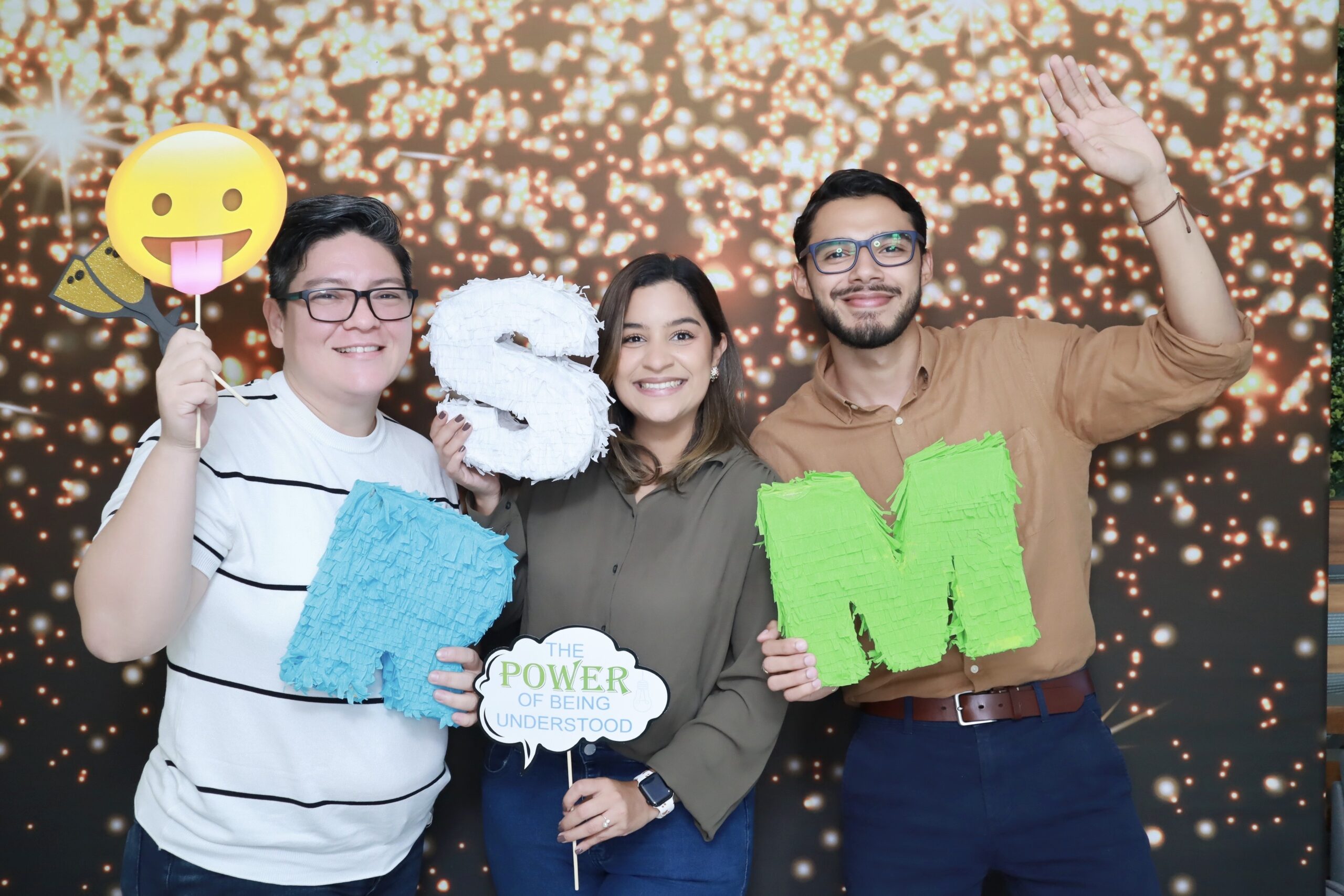 Three people stand smiling in front of a sparkling backdrop. They hold props including emoji, letters "S" and "M," and a sign that says "THE POWER OF BEING UNDERSTOOD." One person is waving.