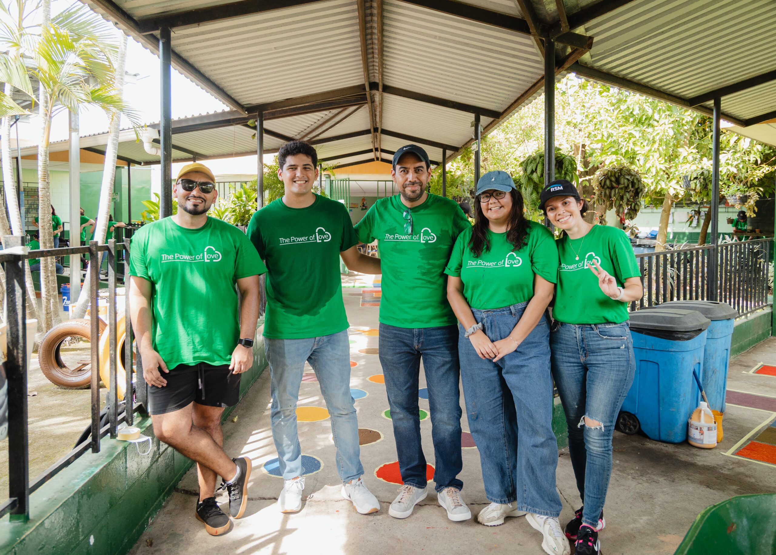 Five people wearing matching green t-shirts stand together smiling under a covered outdoor area, appearing to be part of a volunteer or community event. The background shows trees, painted ground, and playground equipment.