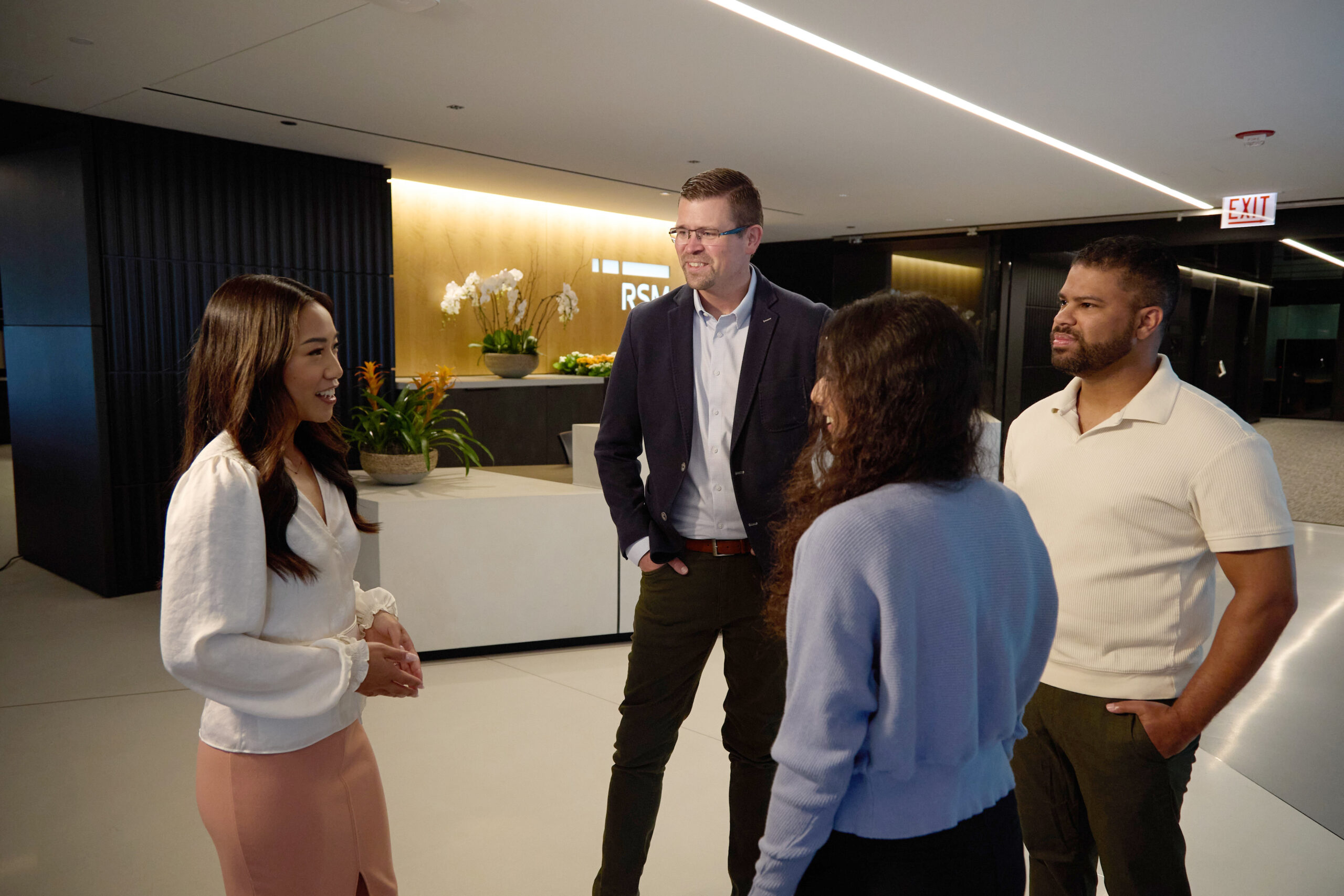 Four people stand in a modern office lobby, engaged in conversation and smiling. There are plants and an illuminated company sign in the background, with a receptionist desk nearby.