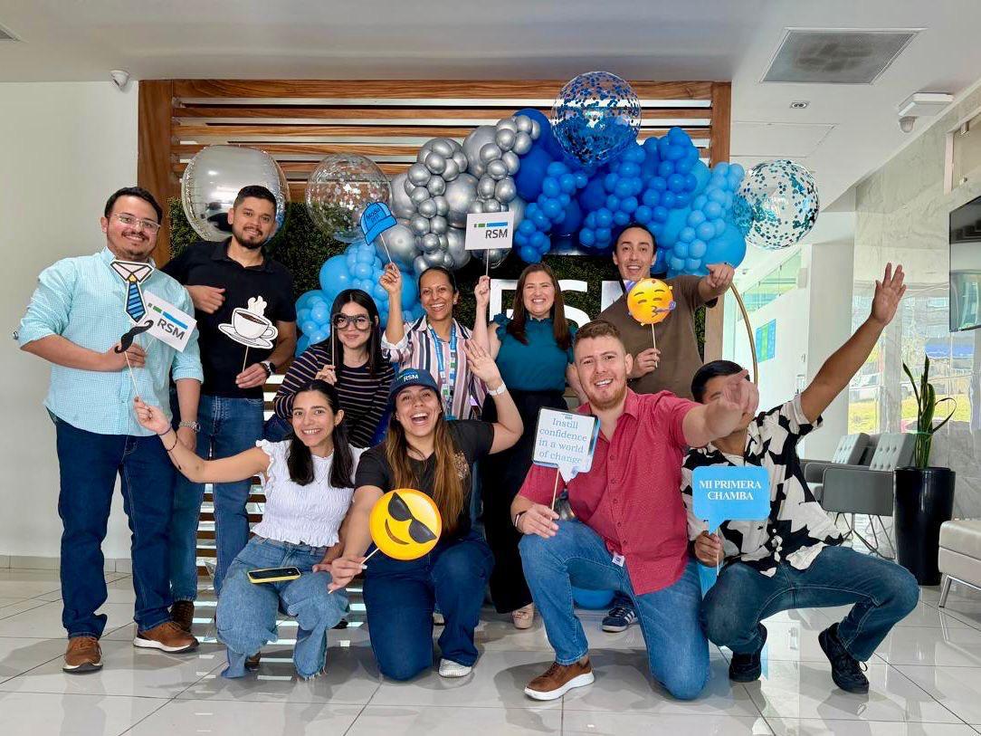 A group of smiling people pose indoors holding fun signs and emoji props in front of a festive blue and silver balloon display. They appear happy and are celebrating together in a modern, bright space.