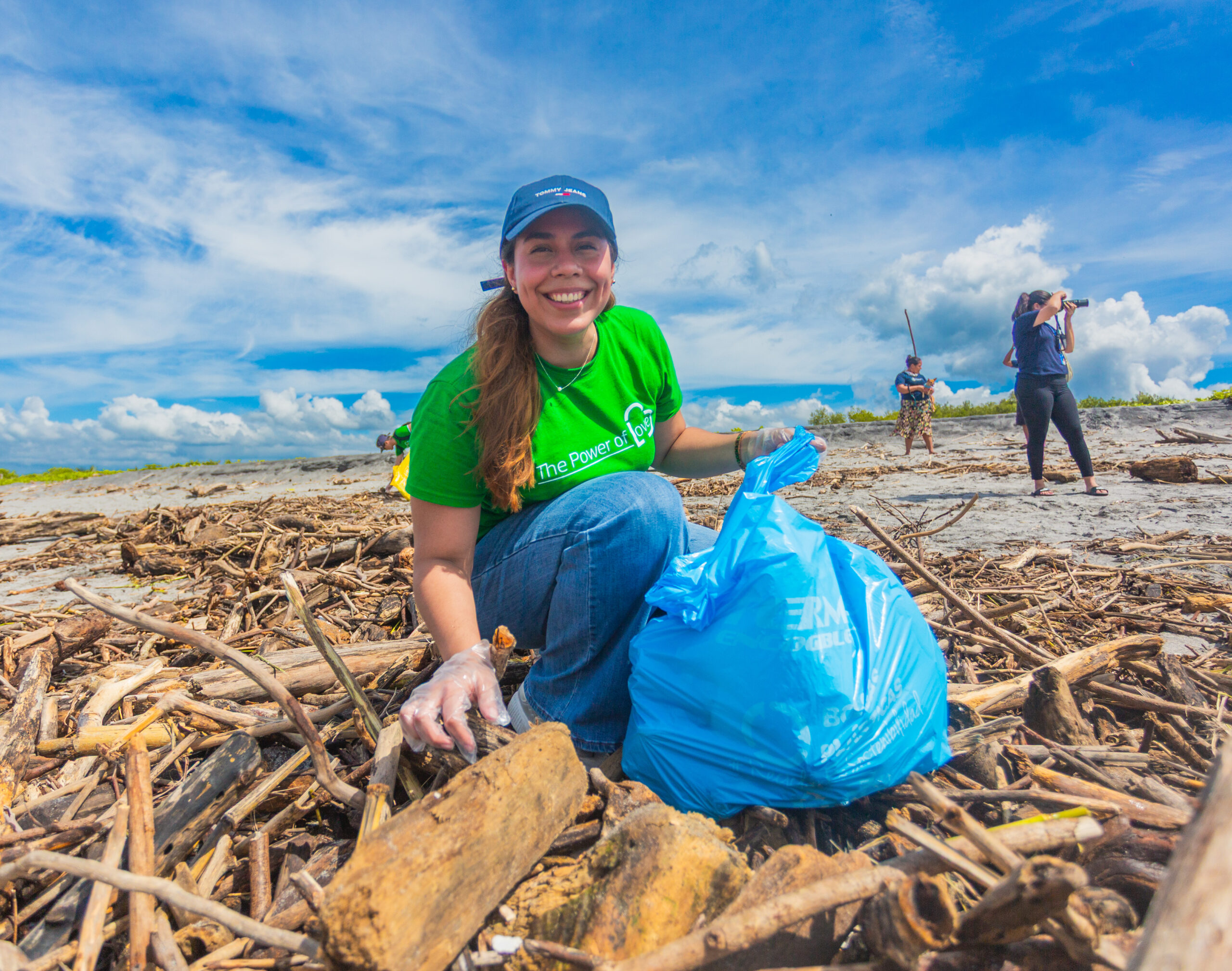 A smiling woman in a green shirt and blue cap picks up litter and driftwood on a beach, kneeling beside a large blue trash bag. Other volunteers are cleaning in the background under a bright, cloudy sky.