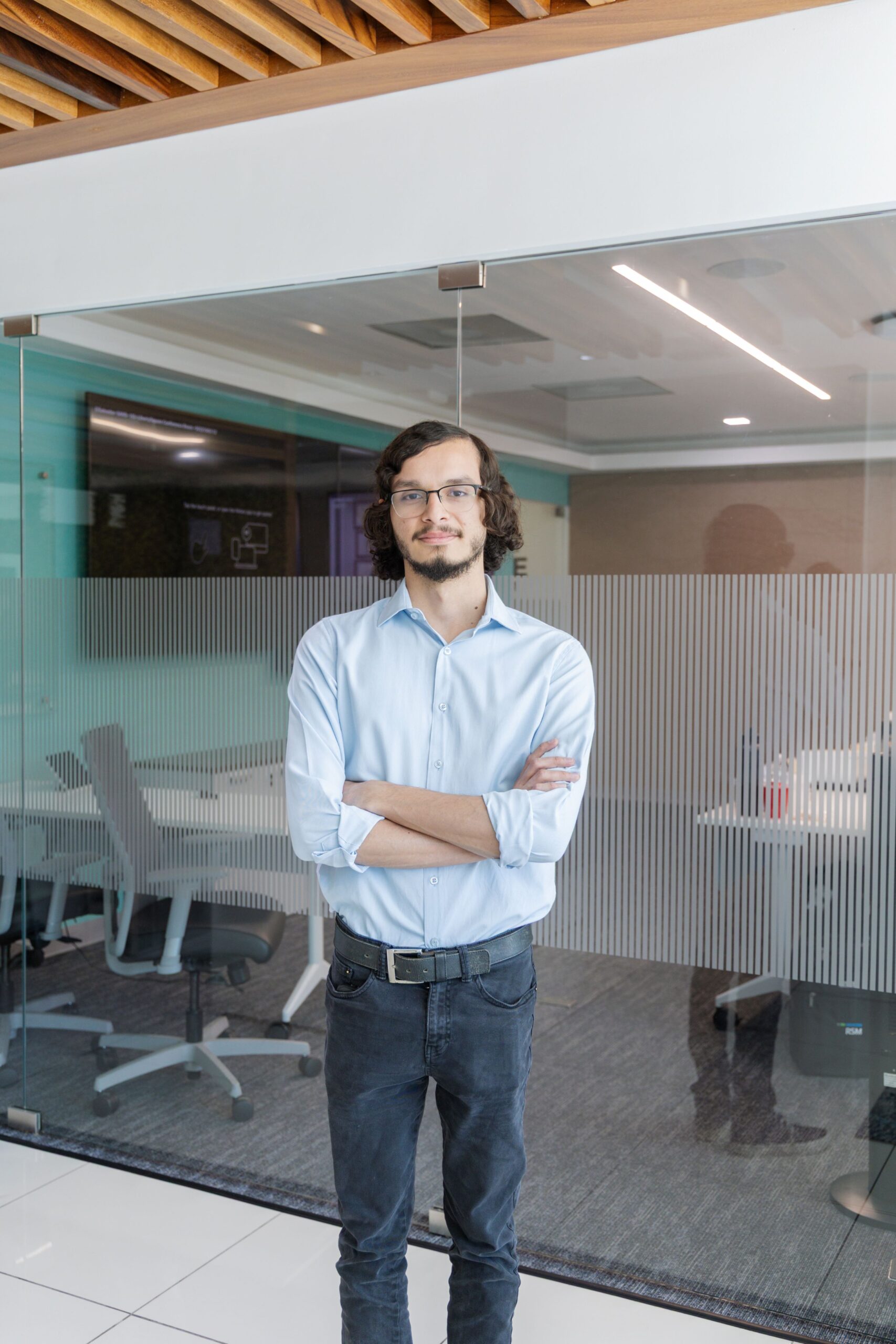 A person with glasses, a light blue shirt, and dark pants stands with arms crossed in a modern office space with glass walls and empty chairs visible inside a meeting room.