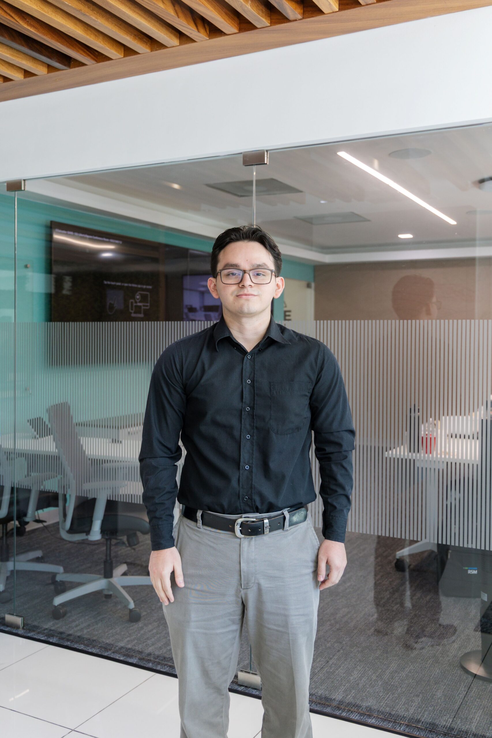 A man wearing glasses, a black shirt, and gray pants stands indoors in front of a glass-walled office meeting room with modern furniture and ceiling panels visible.