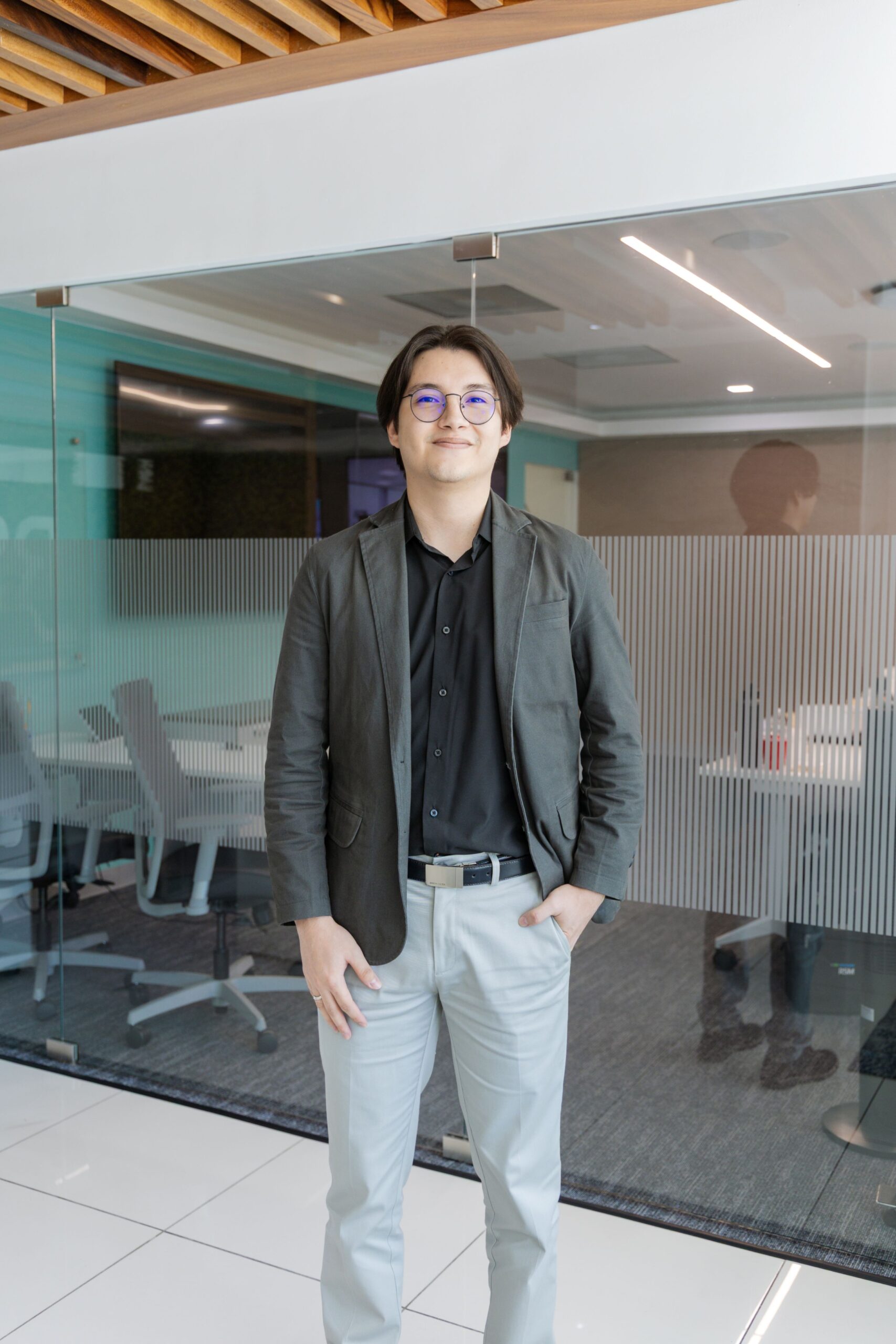 A person with medium-length dark hair, wearing glasses, a dark blazer, black shirt, and light-colored pants, stands and smiles in a modern office space with glass walls and a conference room in the background.
