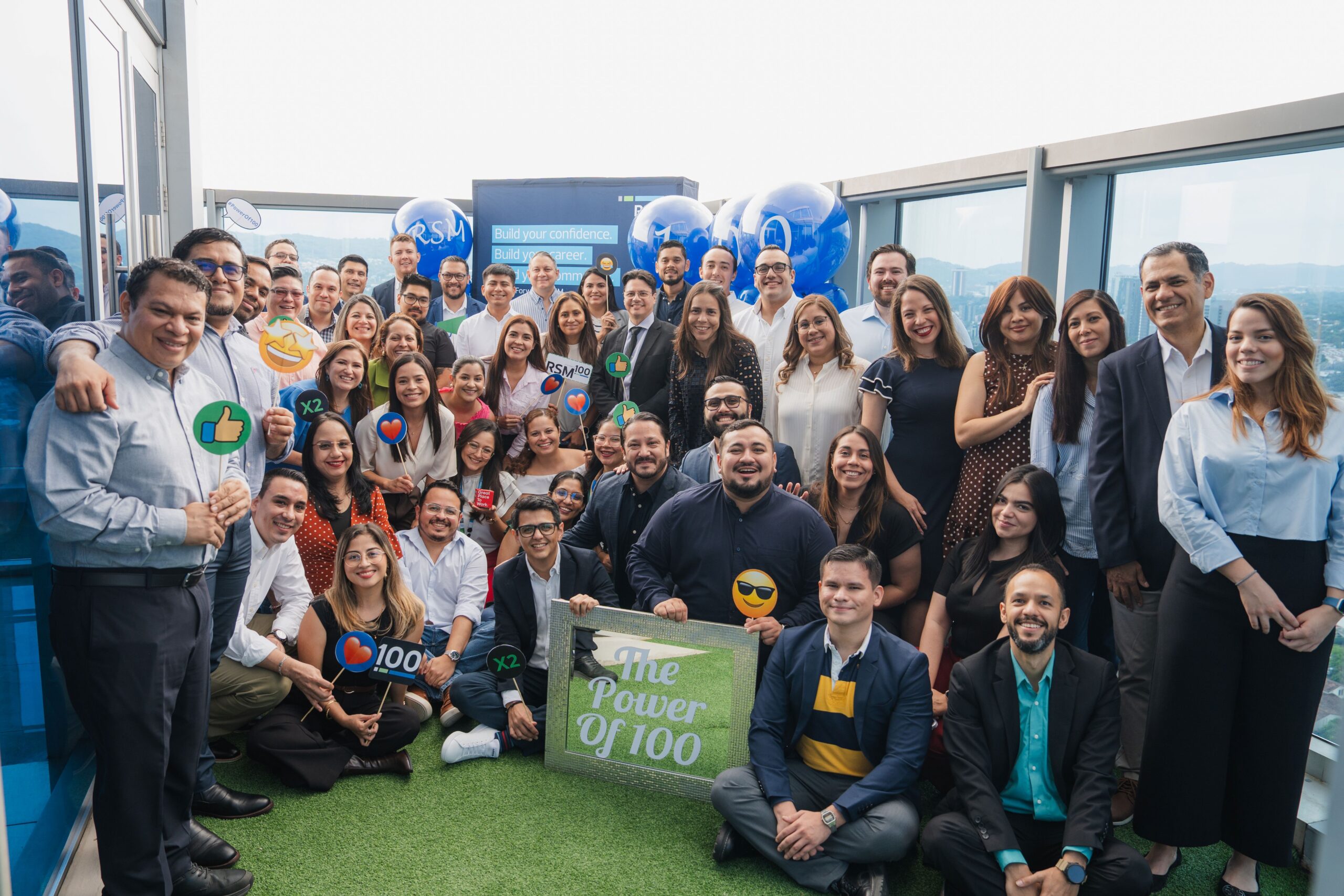 A large group of people smiling and posing on a rooftop with balloons and signs, including one that reads “The Power of 100.” Some hold emoji props. It is daytime, and the mood is celebratory.