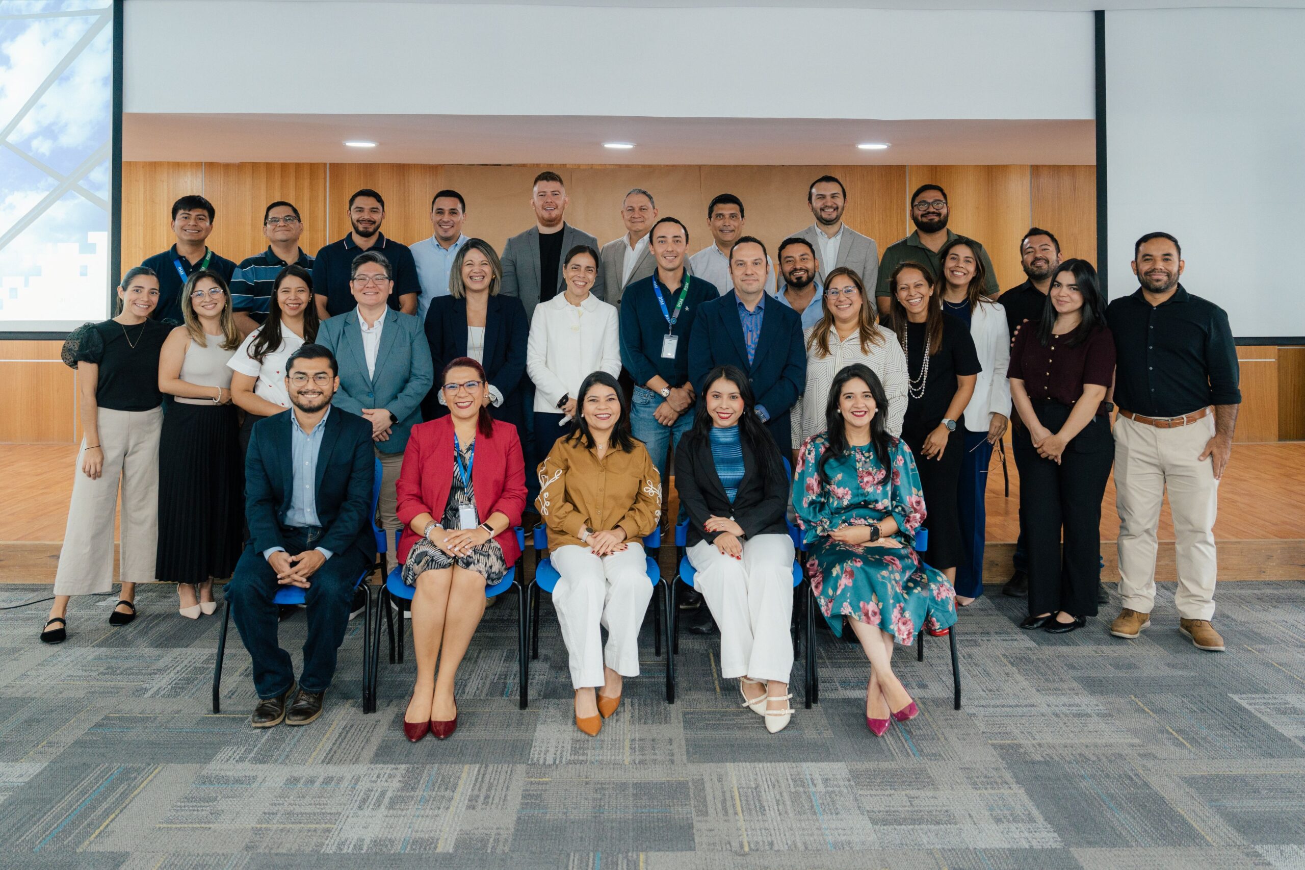 A group of 27 professionally dressed people pose and smile for a group photo in a modern conference room with wooden accents and a projector screen in the background.
