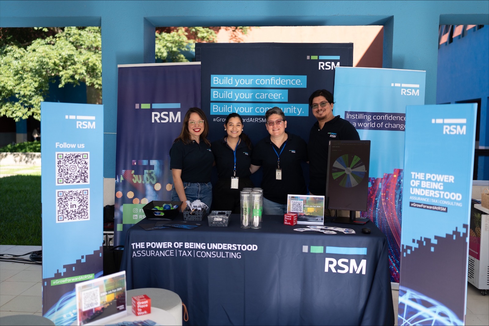 Four people stand smiling behind an RSM booth with banners, promotional items, a QR code sign, and a spinning prize wheel. The display features RSM branding and the slogan "The Power of Being Understood.