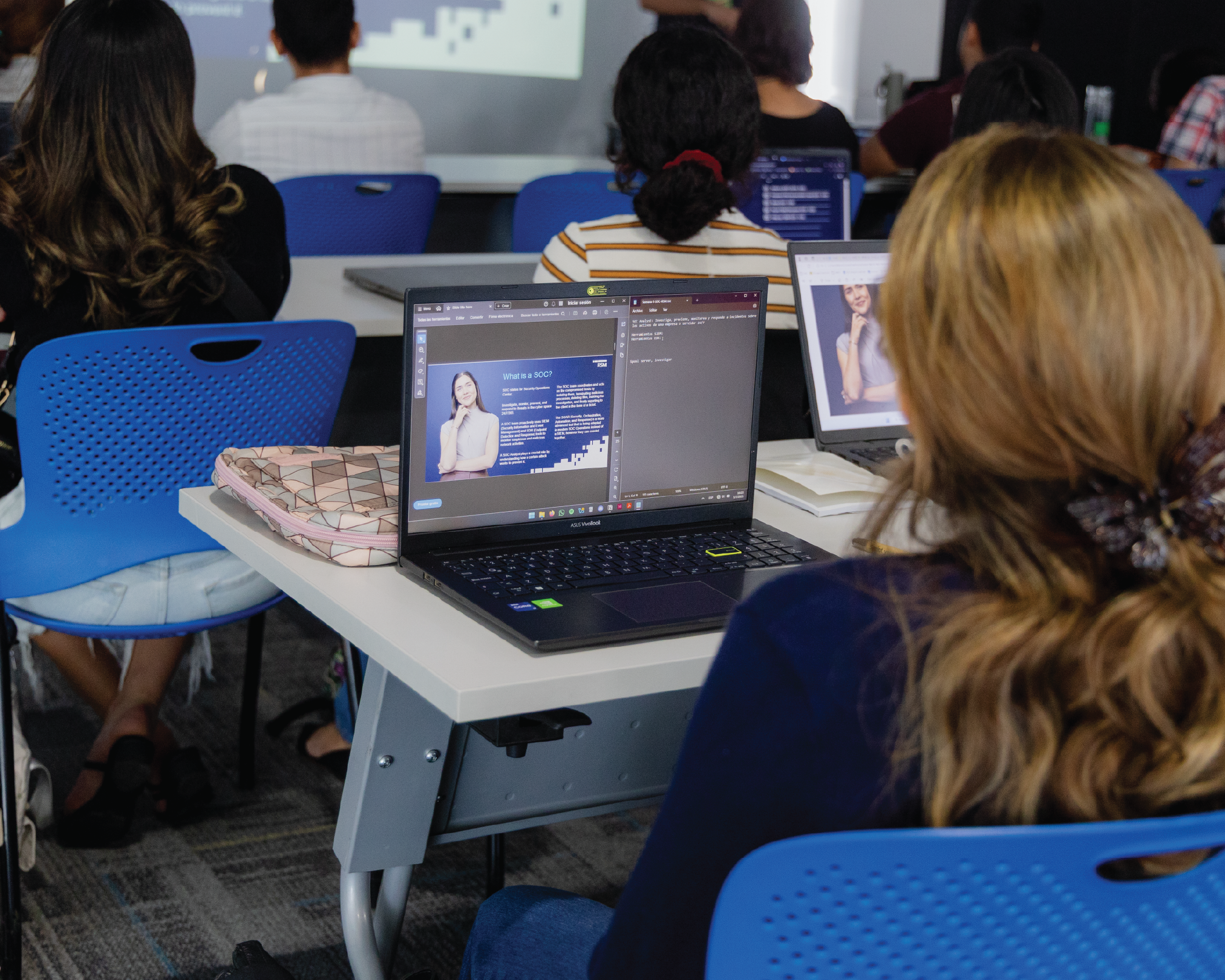 A classroom with students seated at desks, some using laptops. One laptop screen displays a presentation slide with a woman’s photo and text. The focus is on the students’ backs and their workspaces.