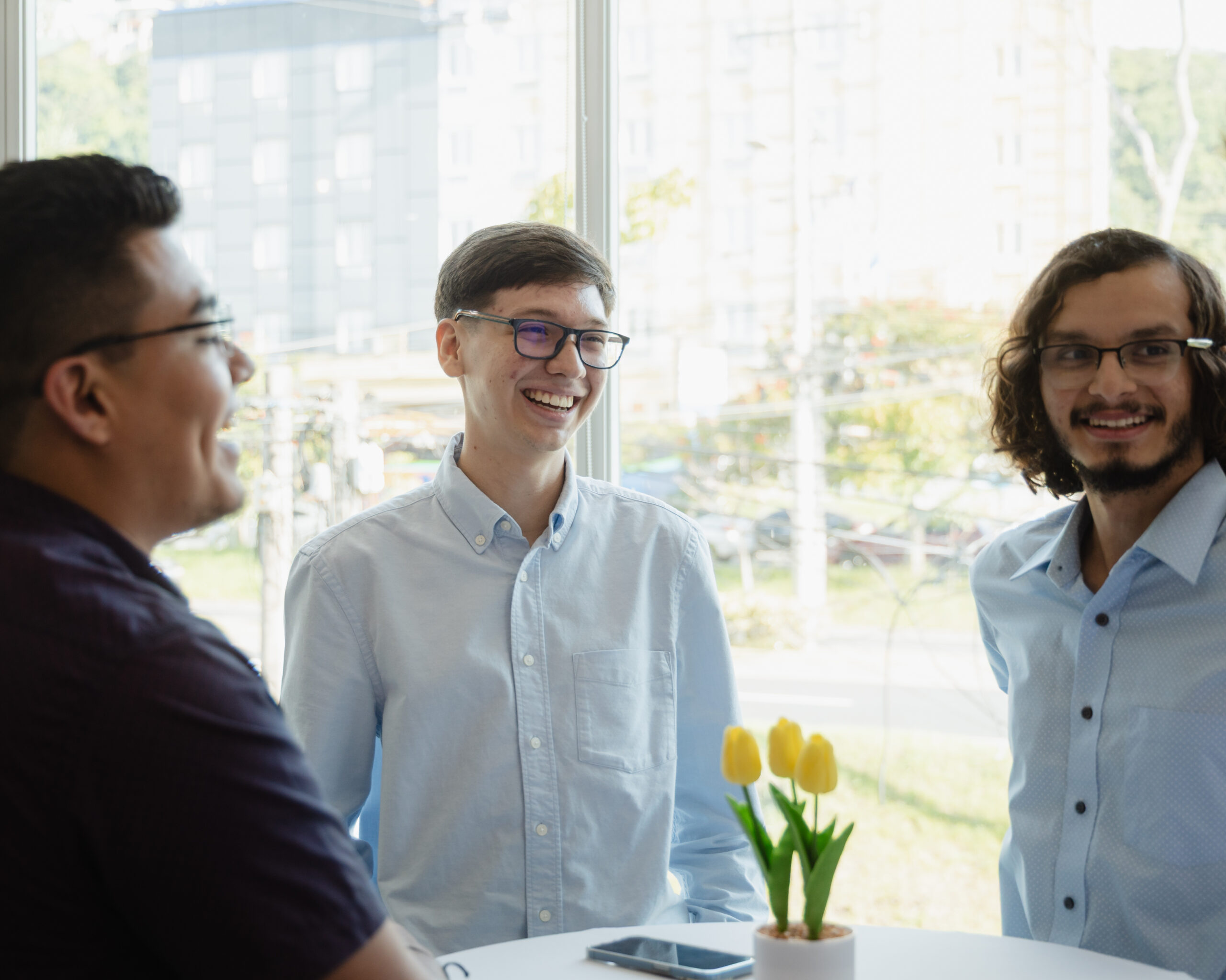 Three men stand and smile around a table with a vase of yellow tulips near a bright window. A smartphone lies on the table in front of them. City buildings and greenery are visible outside.